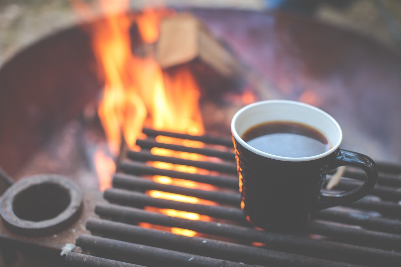 A black cup of coffee next to an open fire on a black rack while camping outdoors.